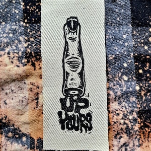 May include: A black and white embroidered patch with a finger pointing upwards and the text "UP YOURS" below it.