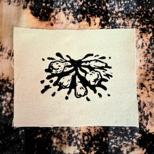 Puede incluir: Un parche de tela blanquecina con un diseño floral negro. El diseño es un grupo estilizado de flores con salpicaduras de tinta. El fondo es una tela vaquera blanqueada.