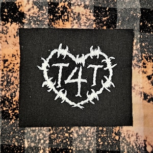Può includere: Toppa in tessuto nero con un design a forma di cuore bianco. Il cuore è circondato da un design a filo spinato. Le lettere "T4T" sono all'interno del cuore.
