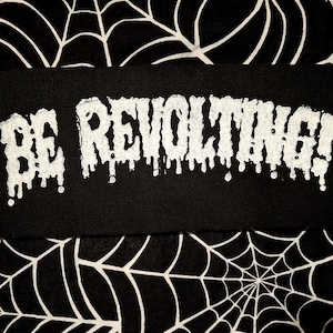 May include: Black rectangular patch with the phrase "BE REVOLTING!" in dripping white letters. The patch is set against a black and white spiderweb pattern.