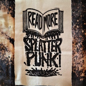 May include: A rectangular patch with the words "READ MORE" above "SPLATTER PUNK!" in a bold, black, distressed font. The design features an open book and dripping ink effects on a beige fabric.