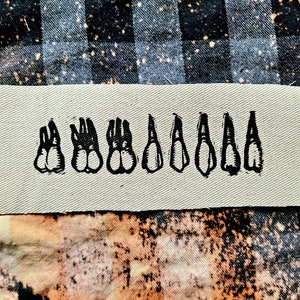 May include: A beige fabric patch with a black embroidered design of eight teeth. The teeth are arranged in a row, with the largest tooth on the left and the smallest tooth on the right.
