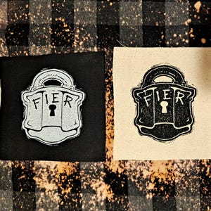 May include: Two fabric patches featuring a black and white image of a padlock with the word "FIER" printed on it. The patches are displayed against a black and white checked background.