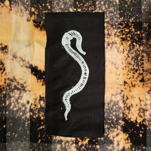 May include: A black patch featuring a white, stylized snake design. The patch is rectangular and set against a backdrop of a plaid pattern in black, tan, and white.