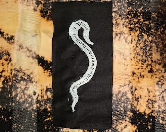 Peut inclure: Un écusson noir avec un motif de serpent blanc stylisé. L'écusson est rectangulaire et placé sur un fond de motif à carreaux noir, beige et blanc.