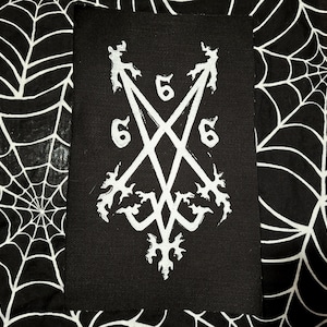 May include: Black rectangular patch with a white inverted pentagram design, featuring the numbers "6" and "5". The patch has a gothic aesthetic with decorative elements. The background is a black and white spiderweb pattern.