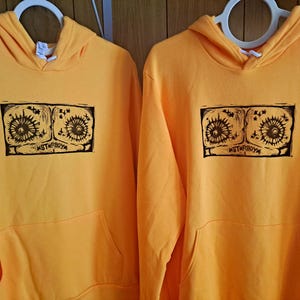 Puede incluir: Dos sudaderas con capucha amarillas con un estampado gráfico negro de dos ojos con el texto "STARBOYZ" debajo.