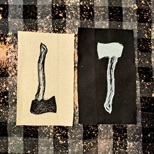 May include: Two fabric patches with an axe design. One patch is beige with a black axe, and the other patch is black with a white axe.