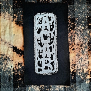 May include: Black fabric patch with a white embroidered design of a stylized abstract shape. The shape is made up of multiple overlapping circles and curves.