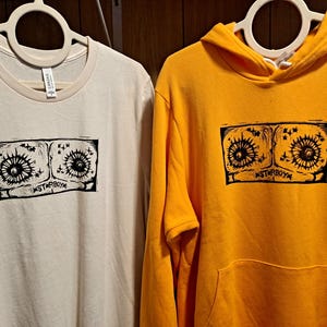May include: Cream-colored t-shirt and yellow hoodie with a black graphic print. The print features two stylized eye-like shapes with radiating lines and the text "STARBOY". Both garments are hanging on white hangers.