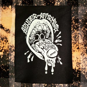 May include: Black rectangular patch featuring a white illustration of an open mouth with the words "GENDER AFFIRMING" and "WHORE MOANS". The patch is set against a plaid background with bleach-like spots.