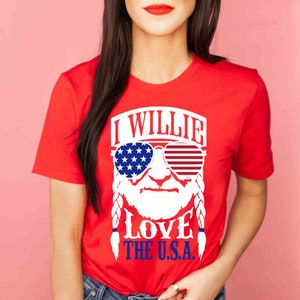 May include: Red t-shirt with a white graphic of Willie Nelson wearing sunglasses with the American flag on them. The text "I Willie Love The U.S.A." is printed on the shirt.