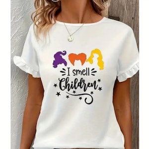 May include: White t-shirt with a graphic of three witches from the movie Hocus Pocus. The text "I smell children" is printed below the witches.