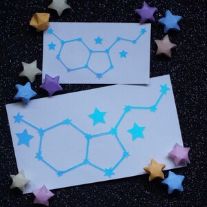 May include: Two white cards with blue constellation outlines of the Gemini zodiac sign. The cards are surrounded by origami stars in various colors, including yellow, purple, and pink, on a black sparkly background.