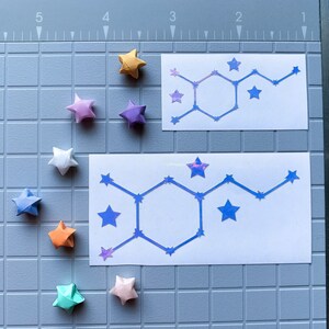 Pode incluir: Vista superior de estrelas de origami coloridas, incluindo amarelo, roxo, branco, azul, laranja e verde. Dois adesivos retangulares brancos com um design de estrelas azuis. Fundo: uma base de corte cinza.