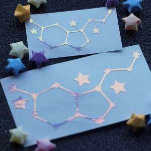 May include: Two blue cards with a white iridescent outline of a chemical structure, resembling a molecule, on each card. The cards are surrounded by origami stars in various colors, including yellow, blue, and purple.