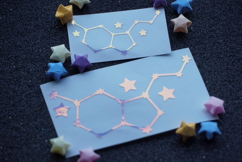 May include: Two blue cards with a white iridescent outline of a chemical structure, resembling a molecule, on each card. The cards are surrounded by origami stars in various colors, including yellow, blue, and purple.