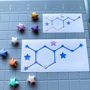 Pode incluir: Uma vista a&eacute;rea mostra estrelas de origami multicoloridas, incluindo azul, laranja e rosa, espalhadas em torno de dois adesivos brancos. Os adesivos apresentam um design geom&eacute;trico com um padr&atilde;o de constela&ccedil;&atilde;o em azul e roxo iridescente.