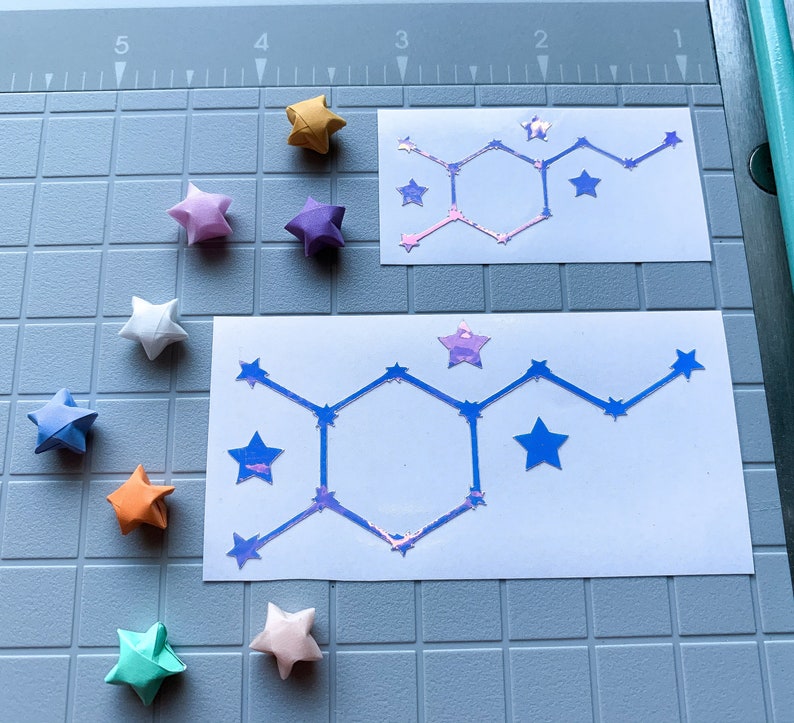 Pode incluir: Uma vista a&eacute;rea mostra estrelas de origami multicoloridas, incluindo azul, laranja e rosa, espalhadas em torno de dois adesivos brancos. Os adesivos apresentam um design geom&eacute;trico com um padr&atilde;o de constela&ccedil;&atilde;o em azul e roxo iridescente.