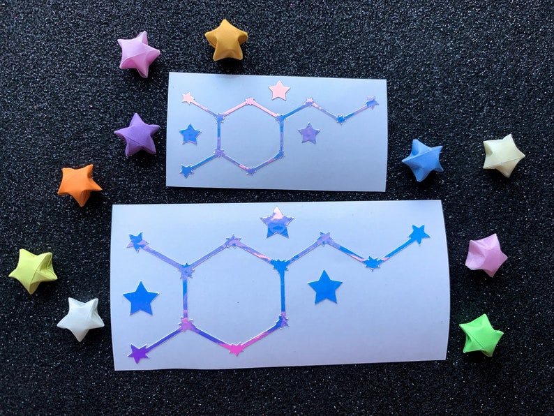 Pode incluir: Dois adesivos brancos com um design hologr&aacute;fico de estrutura molecular e estrelas. Ao redor, estrelas de origami coloridas em tons de rosa, amarelo, laranja, azul e verde.