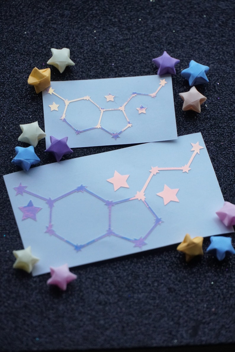 May include: Two blue cards with a constellation design made of iridescent stars. The cards are surrounded by origami stars in various colors, including yellow, pink, blue, and purple.