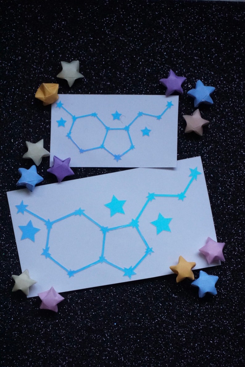 May include: Two white cards with blue constellation outlines of the Gemini zodiac sign. The cards are surrounded by origami stars in various colors, including yellow, purple, and pink, on a black sparkly background.
