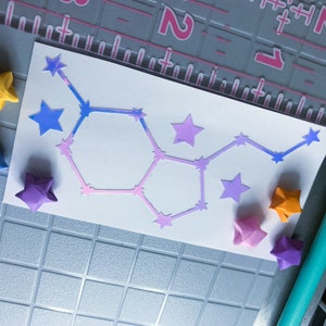 May include: A white card with a pink and blue constellation design. The card is surrounded by origami stars in various colors, including yellow, blue, purple, and orange.