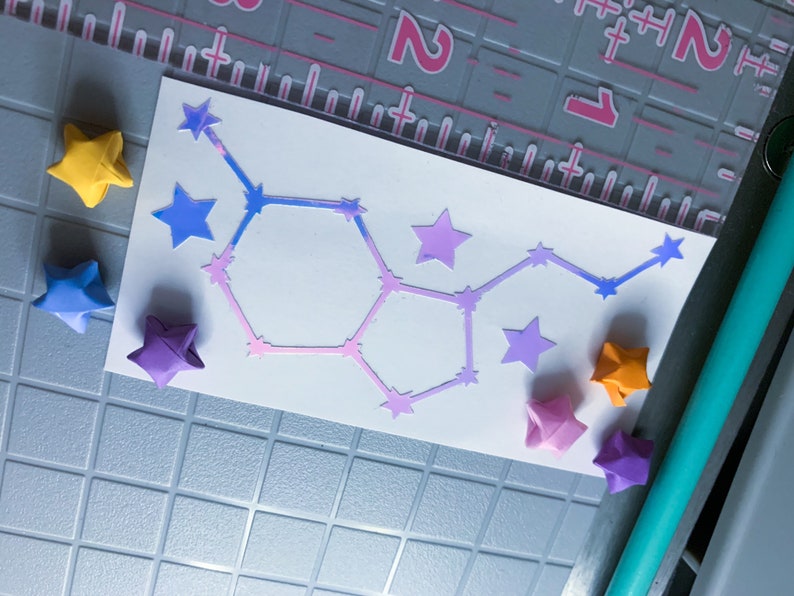 May include: A white card with a pink and blue constellation design. The card is surrounded by origami stars in various colors, including yellow, blue, purple, and orange.