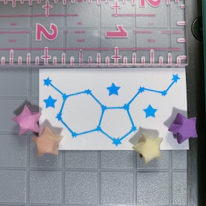 May include: A white card with a blue constellation design and five origami stars in pink, light pink, and purple.