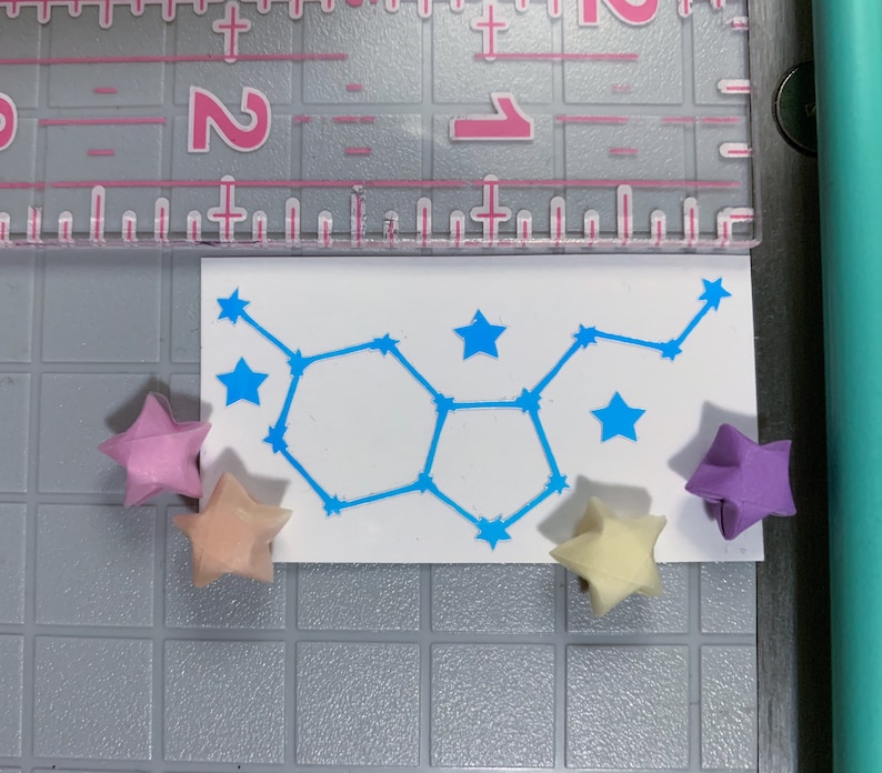 May include: A white card with a blue constellation design and five origami stars in pink, light pink, and purple.