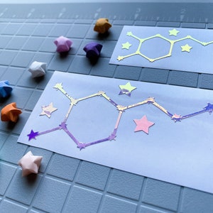 Pode incluir: Duas etiquetas retangulares brancas com um design de estrutura molecular e detalhes de estrelas. As etiquetas t&ecirc;m um efeito hologr&aacute;fico, refletindo v&aacute;rias cores. Estrelas de origami multicoloridas est&atilde;o espalhadas.