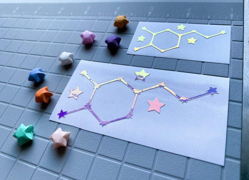 Pode incluir: Duas etiquetas retangulares brancas com um design de estrutura molecular e detalhes de estrelas. As etiquetas t&ecirc;m um efeito hologr&aacute;fico, refletindo v&aacute;rias cores. Estrelas de origami multicoloridas est&atilde;o espalhadas.