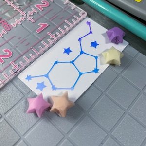 May include: A white card with a blue constellation design and five origami stars in pink, purple, and cream colors. The stars are arranged around the card.