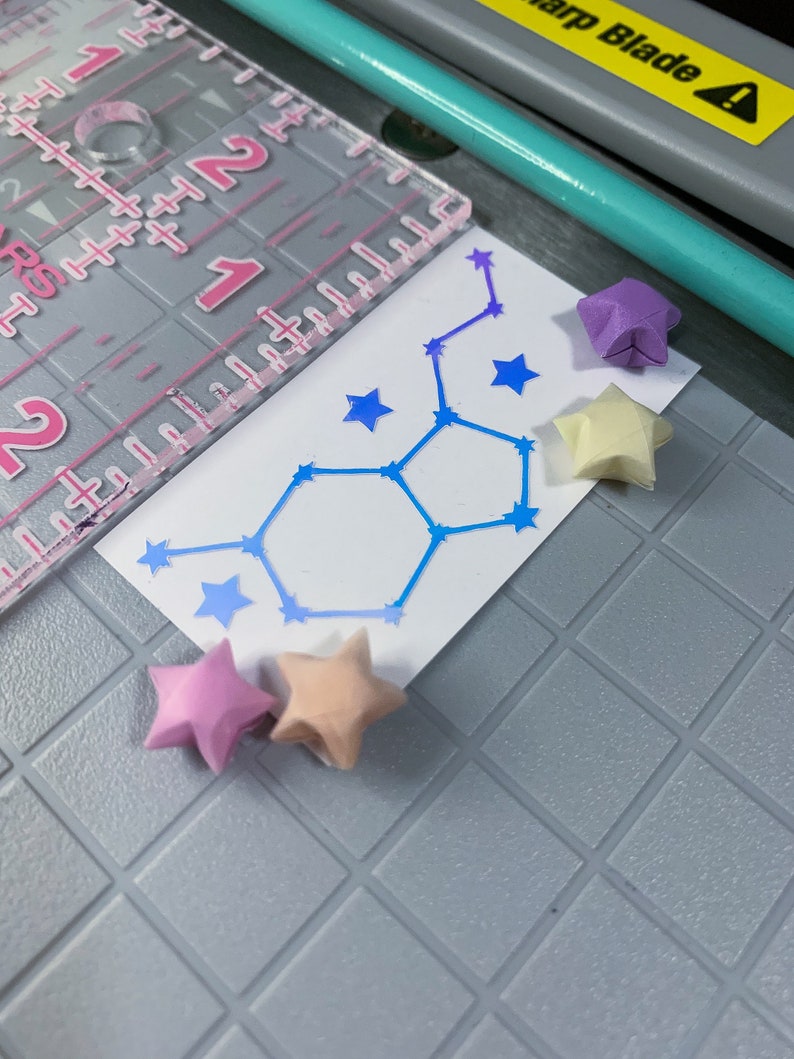 May include: A white card with a blue constellation design and five origami stars in pink, purple, and cream colors. The stars are arranged around the card.