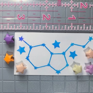 May include: A white card with a blue constellation design. The constellation is surrounded by origami stars in various colors, including purple, orange, peach, and light pink.