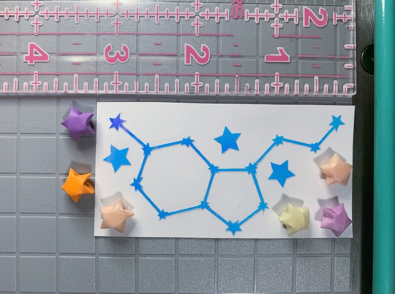 May include: A white card with a blue constellation design. The constellation is surrounded by origami stars in various colors, including purple, orange, peach, and light pink.