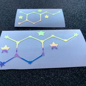 Pode incluir: Dois autocolantes hologr&aacute;ficos com um design geom&eacute;trico, um hex&aacute;gono central e detalhes de estrelas. Os autocolantes t&ecirc;m um gradiente de cores, incluindo amarelo, azul e roxo. Os autocolantes est&atilde;o num fundo cinzento.