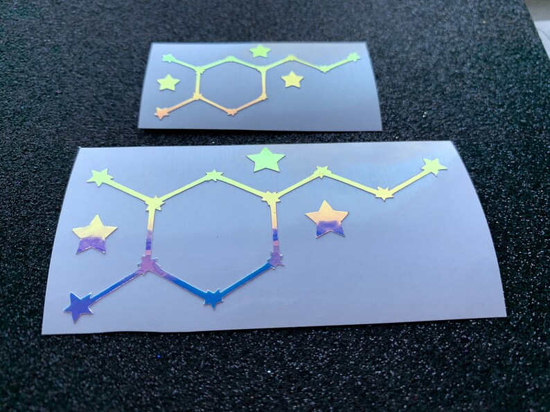 Pode incluir: Dois autocolantes hologr&aacute;ficos com um design geom&eacute;trico, um hex&aacute;gono central e detalhes de estrelas. Os autocolantes t&ecirc;m um gradiente de cores, incluindo amarelo, azul e roxo. Os autocolantes est&atilde;o num fundo cinzento.