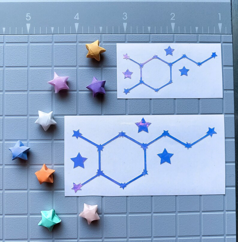 Pode incluir: Vista superior de estrelas de origami coloridas, incluindo amarelo, roxo, branco, azul, laranja e verde. Dois adesivos retangulares brancos com um design de estrelas azuis. Fundo: uma base de corte cinza.