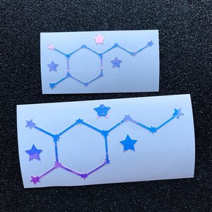 Pode incluir: Dois adesivos retangulares brancos com um design hologr&aacute;fico. Cada adesivo apresenta uma forma geom&eacute;trica com detalhes de estrelas em tons de azul, roxo e rosa. Os adesivos s&atilde;o exibidos em uma superf&iacute;cie preta texturizada.