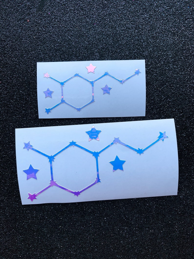 Pode incluir: Dois adesivos retangulares brancos com um design hologr&aacute;fico. Cada adesivo apresenta uma forma geom&eacute;trica com detalhes de estrelas em tons de azul, roxo e rosa. Os adesivos s&atilde;o exibidos em uma superf&iacute;cie preta texturizada.