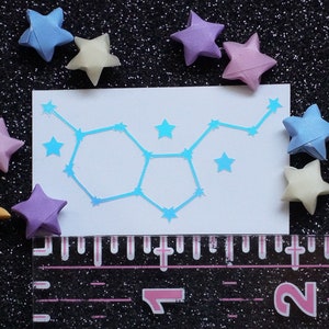 May include: A white card with a blue iridescent constellation design. The constellation is of the Gemini zodiac sign. The card is surrounded by origami stars in various colors, including pink, blue, yellow, and purple.