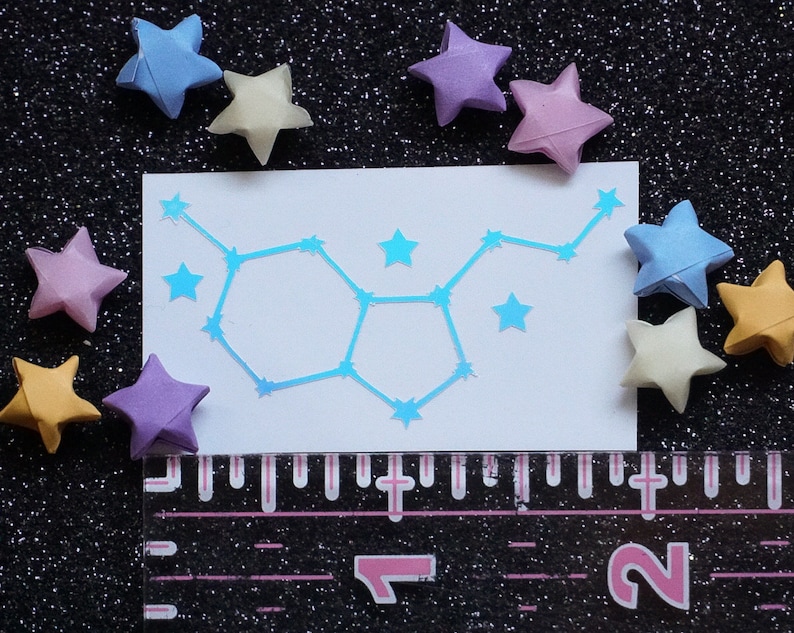 May include: A white card with a blue iridescent constellation design. The constellation is of the Gemini zodiac sign. The card is surrounded by origami stars in various colors, including pink, blue, yellow, and purple.