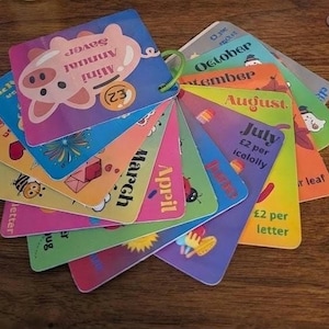 May include: A fan-shaped set of colourful laminated cards held together by a green plastic ring. The top card features a pink pig illustration with the text Mini Annual Saver and a circle with the number 2. Other cards display months like August, July, and March with various seasonal graphics and text.