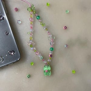 Może przedstawiać: Różowo-zielony pasek do telefonu z zielonym gumowym misiem. Pasek wykonany jest z małych plastikowych koralików w kształcie gwiazd, serc i kwiatów.