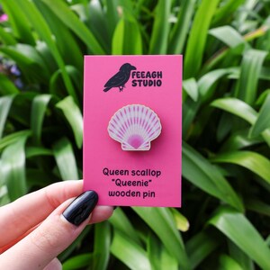 May include: A pink card with a black raven logo and the text "FEEAGH STUDIO" displays a wooden pin. The pin is a scallop shell design with white and pink stripes. The text "Queen scallop 'Queenie' wooden pin" is below.