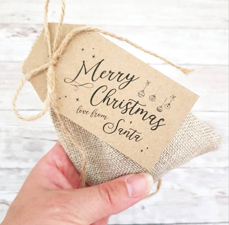 May include: A small burlap gift bag tied with twine, with a brown paper tag. The tag says "Merry Christmas love from Santa" in elegant script, with star and bauble illustrations. The bag is held against a light wood background.