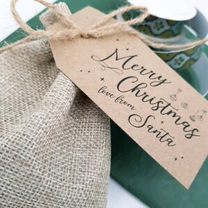 May include: A hessian gift bag tied with twine, with a brown tag. The tag says "Merry Christmas, love from Santa" in cursive, with star and bauble illustrations. A green surface and a decorative cup are in the background.