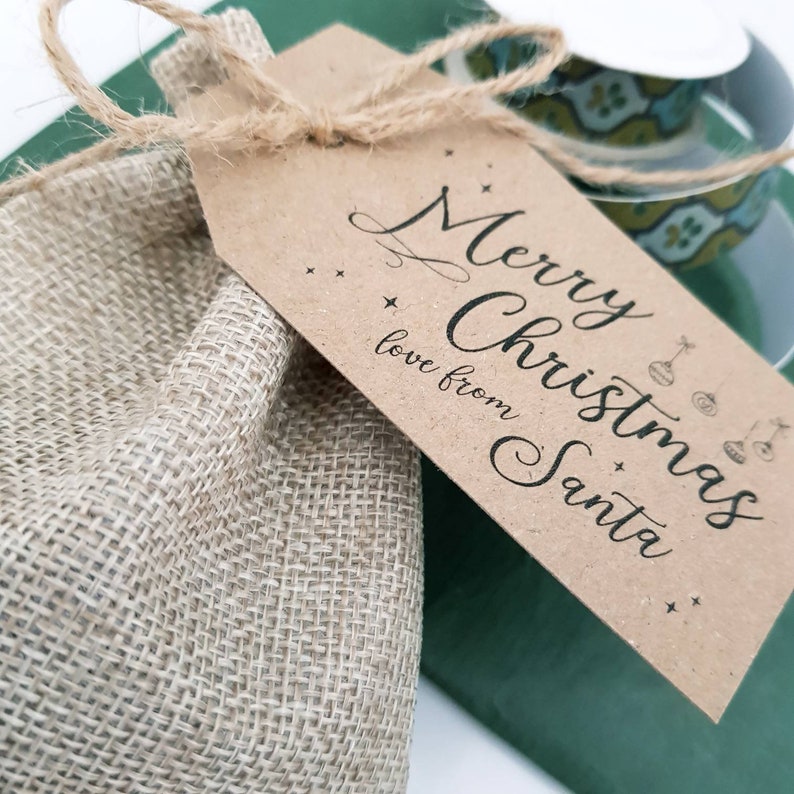 May include: A hessian gift bag tied with twine, with a brown tag. The tag says "Merry Christmas, love from Santa" in cursive, with star and bauble illustrations. A green surface and a decorative cup are in the background.