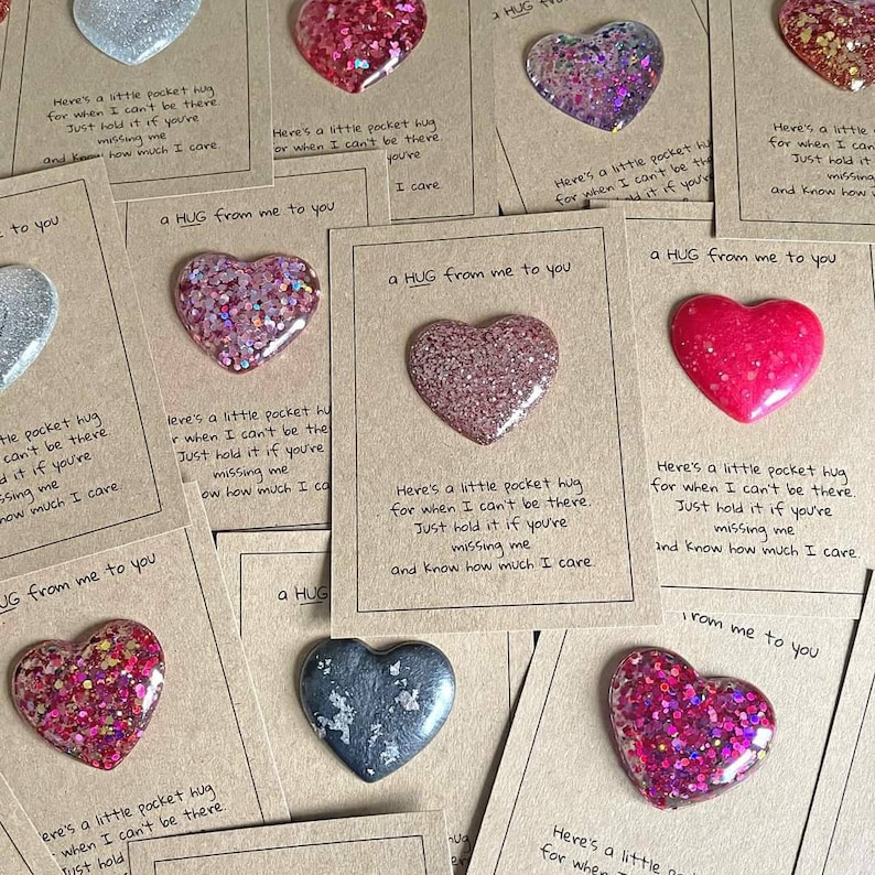 May include: A set of eight small cards with a heart-shaped resin charm attached to each. The charms are filled with glitter and come in a variety of colors, including pink, red, silver, and gray. Each card has a message that reads "a hug from me to you" and includes a short message about missing someone.